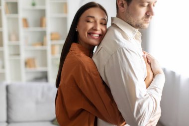 Mutlu, sevgi dolu bir kadın kocasına arkadan sarılıyor ve gülümsüyor. Oturma odasının iç tarafında duruyor. İlişkilerde sevgi ve şefkat