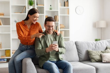 Akıllı telefon kullanan mutlu Avrupalı bir çift, cep telefonu ekranına bakan bir eş, oturma odasındaki koltukta oturan bir eş, boş alan.