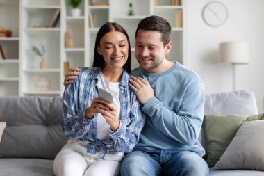 Mutlu genç Avrupalı çift koltukta otururken cep telefonu kullanıyor, kadın kocasıyla içeriği paylaşıyor, yeni başvurular gösteriyor