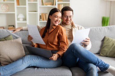 Evrakları ve dizüstü bilgisayarı olan mutlu eşler, tartışma ve finansal planlama ile meşgul, rahat oturma odasında kanepede oturuyor.