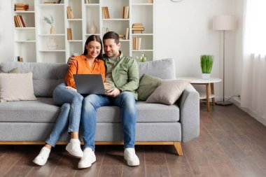 Pozitif genç eşler rahat bir şekilde kanepede oturuyor, gülümseyerek dizüstü ekrana bakıyor, rahat oturma odasında dinleniyor, fotokopi odası, tam boy