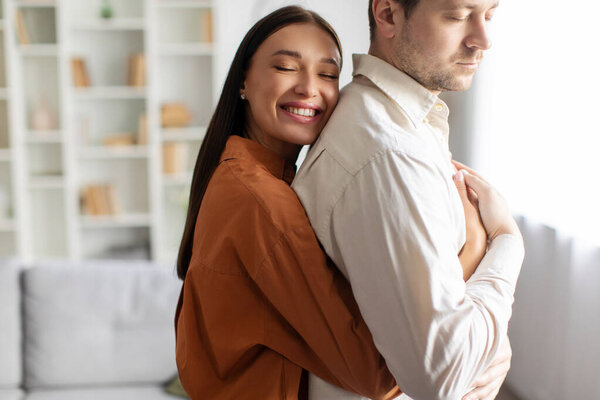 Счастливая любящая женщина обнимает мужа со спины и улыбается, стоя в уютном интерьере гостиной. Любовь и нежность в отношениях