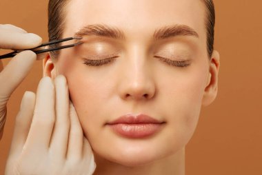 Kozmetik uzmanı elinde cımbızla genç Avrupalı bayana kaş şekillendiriyor. Güzellik uzmanı, kaş kılı yoluyor.