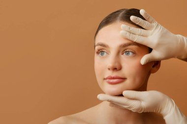 Eldivenli kozmetik uzmanı güzellik ya da estetik ameliyattan önce bayanların yüzüne dokunup kontrol ediyor, bej arka plan üzerinde poz veriyor, serbest alan