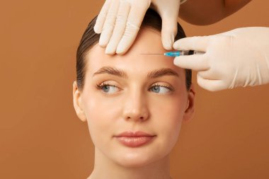 Güzellik tedavileri konsepti. Genç Avrupalı bir bayan güzellik kliniğinde alnına estetik yaptırıyor, bej rengi stüdyo arka planı.