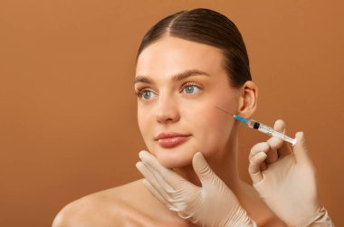 Estetik cerrahi ve kozmetik konsepti. Güzel Avrupalı kadın güzellik iğnesi yaptırıyor, eldivenli kozmetik uzmanı kadın yüzünün yanında şırınga tutuyor, bej arka plan