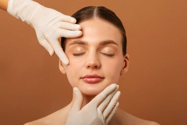 Plastik cerrahi veya güzellik tedavisinden önce kadın yüzüne dokunan kozmetik uzmanı, bej arka planda poz veren kapalı gözlü kadın. Estetik cerrahi ve yüz bakımı konsepti