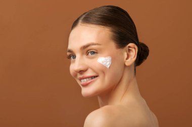 Yanağında güzel bir krem kalp olan genç bir bayan, yeni kozmetik ürünlerin tadını çıkarıyor, kahverengi stüdyo arka planının üzerinde çıplak duruyor.