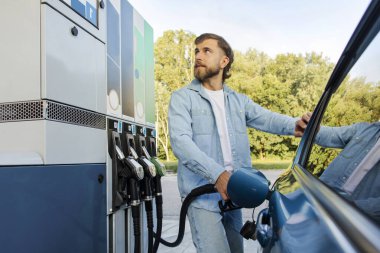 Yakışıklı genç adam benzin istasyonunda duruyor, arabasına yakıt dolduruyor ve benzin sayacına bakıyor.