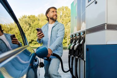 Sakallı adam yakıt ikmali yapıyor ve akıllı telefon kullanıyor, benzin göstergesi ve benzin istasyonu web sitesindeki yakıt fiyatlarını karşılaştırıyor