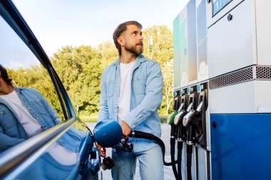 Yakışıklı sakallı Avrupalı adam arabasına yakıt dolduruyor ve skorborda bakıyor, self servis benzin istasyonunda duruyor.