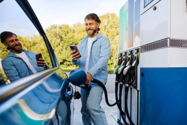 Arabasına yakıt dolduran, akıllı telefondan mesaj atan, otomobili biyo yakıtla dolduran mutlu yakışıklı bir adam.