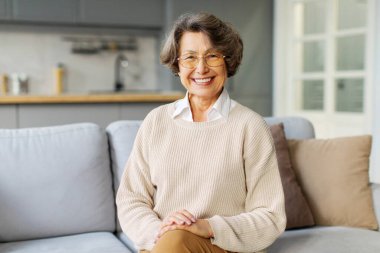 Mutlu, yaşlı bir kadın kameraya gülümsüyor, evde tek başına oturuyor, pozitif, emekli bir kadın oturma odasında rahatlayıp portre için poz veriyor.