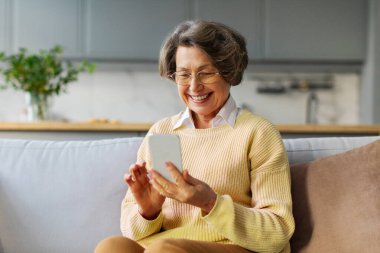 Akıllı telefondan konuşan neşeli yaşlı kadın, online iletişimden zevk alıyor ve gülümsüyor, evdeki kanepede oturuyor, boş yer var.