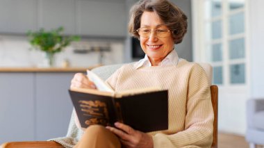 Çekici, neşeli, son sınıf öğrencisi bir kadın sandalyeye oturup kitap okuyor, keyfi yerinde ve hafta sonunun keyfini çıkarıyor.