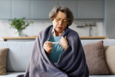 Ateşini ölçen hasta yaşlı bir kadın, kanepeye sarılmış bir şekilde oturuyor, mevsimsel gribi olan yaşlı bir bayan ya da gripten dolayı sağlıksız hisseden soğuk bir kadın.