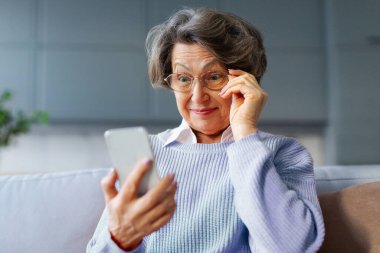 Heyecanlı, şaşırmış bir kadın, güzel haberlerle şoke olmuş, akıllı telefon ekranına geniş gözlerle bakıyor, gözlüklere dokunuyor, kanepede oturuyor.