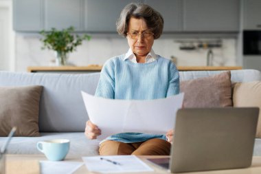 Aile bütçesini dengelemek. Ciddi, yaşlı bir kadın düşünceli bir şekilde kağıt faturalarını okuyor ve dizüstü bilgisayar kullanıyor, internetten finans yönetiyor, evdeki kanepede oturuyor.