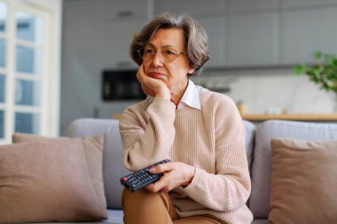 Elinde uzaktan kumandayla evde oturan üzgün yaşlı bir kadın. Sıkılmış, TV 'de izleyecek ilginç bir şeyler arıyor.