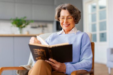 Çekici, neşeli, son sınıf öğrencisi bir kadın sandalyeye oturup kitap okuyor, keyfi yerinde ve hafta sonunun keyfini çıkarıyor.