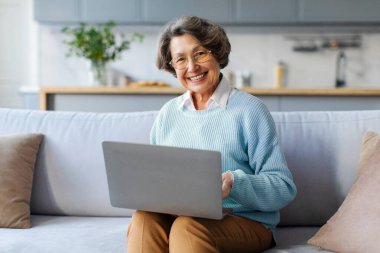 Uzaktan alışveriş için dizüstü bilgisayar kullanan mutlu Avrupalı bir kadın. Evdeki koltukta oturuyor ve kameraya gülümsüyor. Büyükannem serbest çalışıyor.