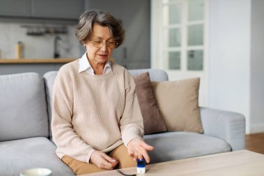 Elinde nabız oksimetresi olan, oksijen doygunluğunu ölçen, evdeki koltukta oturan yaşlı bir kadın. Nabız oksimetresi tıbbi cihaz, emeklilik konseptinde sağlık hizmeti