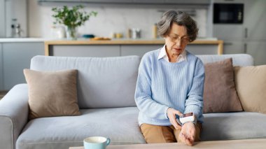 Modern tonometre hipertansiyonu olan yaşlı bir kadın evinde kan basıncını ölçüyor, kanepede oturuyor. Günlük sağlık kontrolleri