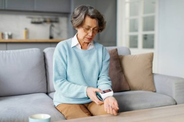 Yaşlı Avrupalı kadın, koltukta otururken sağlık sonuçlarını kontrol etmek için modern tansiyon aleti kullanıyor. Sağlık hizmeti kavramı