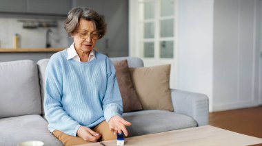 Elinde nabız oksimetresi olan, oksijen doygunluğunu ölçen, evdeki koltukta oturan yaşlı bir kadın. Nabız oksimetresi tıbbi cihaz, emeklilik konseptinde sağlık hizmeti