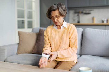 Yaşlı Avrupalı kadın, koltukta otururken sağlık sonuçlarını kontrol etmek için modern tansiyon aleti kullanıyor. Sağlık hizmeti kavramı