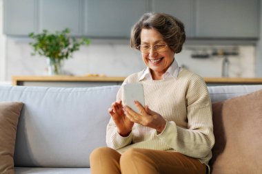 Akıllı telefondan konuşan neşeli yaşlı kadın, online iletişimden zevk alıyor ve gülümsüyor, evdeki kanepede oturuyor, boş yer var.