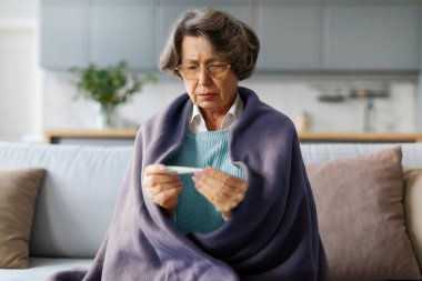 Üst düzey Avrupalı kadın battaniyeye sarılmış termometreye dikkatle bakıyor, kendini kötü hissediyor, evdeki oturma odasında kanepede oturuyor.