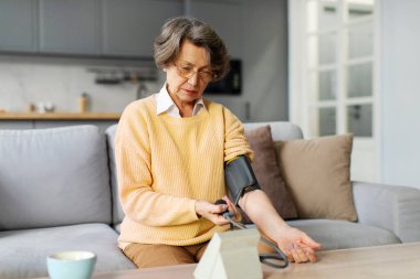 Ciddi kıdemli bir kadın, tansiyon ölçümlerini kontrol etmek için tıbbi cihaz kullanıyor, yüksek tansiyondan muzdarip, evdeki koltukta oturuyor.