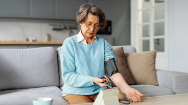 Ciddi kıdemli bir kadın, tansiyon ölçümlerini kontrol etmek için tıbbi cihaz kullanıyor, yüksek tansiyondan muzdarip, evdeki koltukta oturuyor.