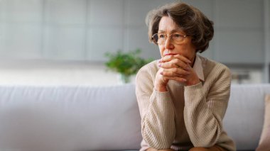Bunalımlı yaşlı bayan çeneye dokunuyor, kanepeye oturuyor ve başka yere bakıyor, endişeli ve stresli hissediyor, sorunları düşünüyor.
