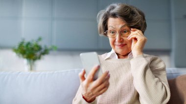 Heyecanlı, şaşırmış bir kadın, güzel haberlerle şoke olmuş, akıllı telefon ekranına geniş gözlerle bakıyor, gözlüklere dokunuyor, kanepede oturuyor.