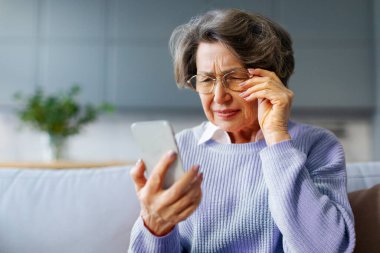 Hayal kırıklığına uğramış yaşlı, gözlüklü bir kadın akıllı telefonuyla sorun yaşıyor, internet bağlantısında sorun yaşıyor, kötü haberler okuyor, kanepede oturuyor.