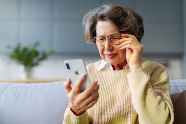 Hayal kırıklığına uğramış yaşlı, gözlüklü bir kadın akıllı telefonuyla sorun yaşıyor, internet bağlantısında sorun yaşıyor, kötü haberler okuyor, kanepede oturuyor.