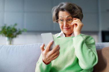 Heyecanlı, şaşırmış bir kadın, güzel haberlerle şoke olmuş, akıllı telefon ekranına geniş gözlerle bakıyor, gözlüklere dokunuyor, kanepede oturuyor.