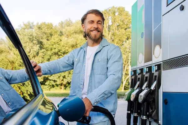 Arabasını yakıtla dolduran mutlu yakışıklı adam modern benzin istasyonunda kameraya gülümseyen bir arabanın yanında duruyor.