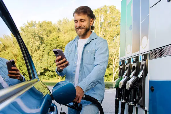 Benzin istasyonunda arabasını benzinle dolduran ve cep telefonu kullanan mutlu bir adam. Telefondaki uygulamayı kullanarak internet üzerinden ödeme yapıyor.