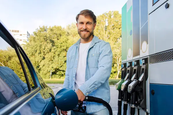 Çevre dostu yakıt ikmali konsepti. Modern benzin istasyonunda arabasını dolduran yakışıklı sakallı adam kameraya gülümsüyor.