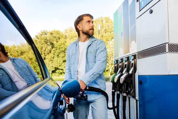 Yakışıklı sakallı Avrupalı adam arabasına yakıt dolduruyor ve skorborda bakıyor, self servis benzin istasyonunda duruyor.