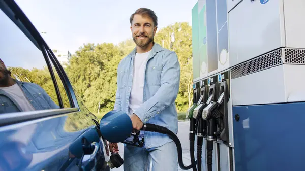 Modern benzin istasyonunda biyoyakıt dolduran yakışıklı bir adam kameraya gülümseyen bir erkek sürücü, panorama çekimi. Otomobil yakıtlama teklifi