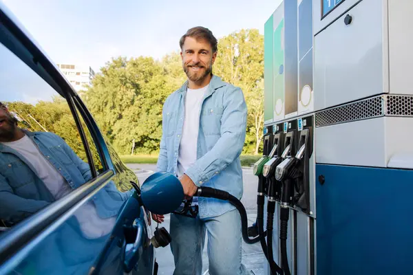 Benzin istasyonunda yakıt ikmali yapan mutlu Avrupalı adam, otomobilin yanında duruyor ve yakıt hortumunu tutuyor, kameraya bakıyor ve gülümsüyor.
