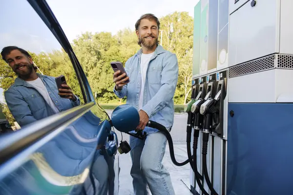 Arabasına yakıt dolduran, benzin istasyonunda cep telefonu kullanan, benzin deposuna benzin döken, kameraya gülümseyen yakışıklı bir adam.