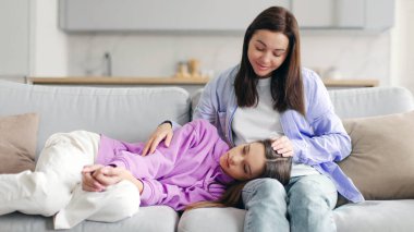 Sevgi dolu bir anne, genç kızıyla konuşuyor, yorgun üzgün bir kız, anne dizlerinin üzerinde yatıyor, evdeki kanepede oturuyor.