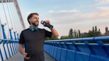 Köprüde duran ve şişeden su içen sporcu, dışarıda antrenman yaparken mola veren spor giyim ve kulaklık takan adam, panorama, bedava alan.