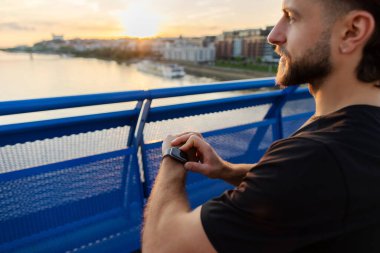 Genç sporcu adam antrenmana ara veriyor. Akıllı fitness saati kullanıyor ve şehir köprüsünden manzaraya bakıyor.