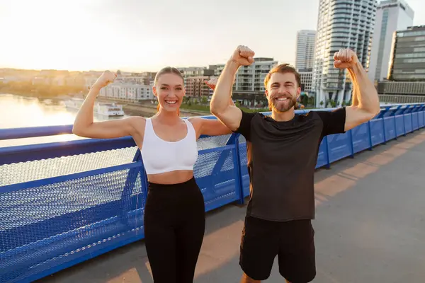 Spor yaparken bisepslerini gösteren mutlu bir erkek ve kadın. Eğitimden sonra şehir köprüsünde poz veriyorlar.